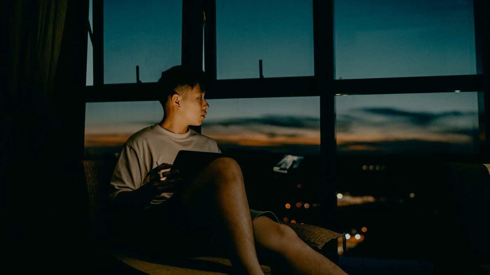 A person sits in a wicker armchair by a large window at dusk, a tablet on their lap.